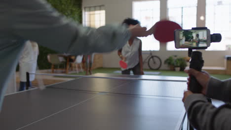 Jóvenes-Empresarios-Jugando-Ping-Pong-En-La-Oficina-Disfrutando-De-Un-Colega-Competitivo-Y-Divertido-Usando-Un-Juego-Para-Compartir-Teléfonos-Inteligentes-En-Las-Redes-Sociales