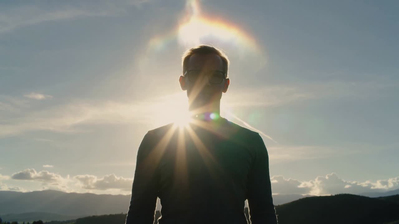Standing man on hilltop, sun aligning near chest creating starburst halo, revealing eyeglasses