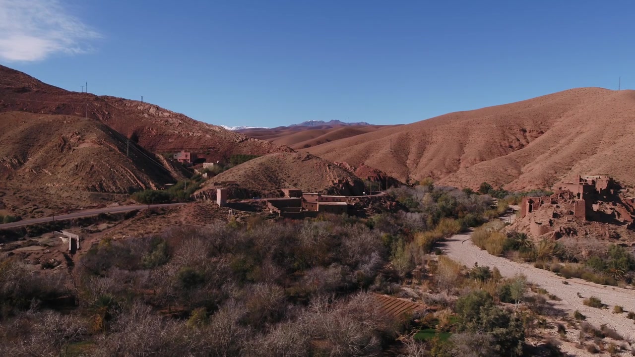 High-angle panoramic aerial drone shot capturing the expansive green oasis, traditional mud-brick Kasbahs, and surrounding red hills in Dades Valley, Morocco.