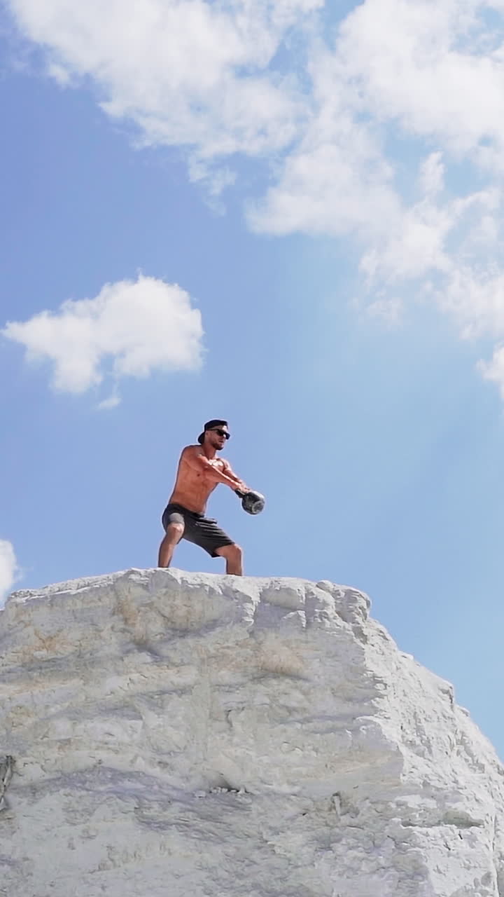 Shirtless athlete lifting kettlebell. Healthy sportsman is swinging kettlebell with two hands on the top of white hill. View from below on bodybuilder under blue sky. Vertical video