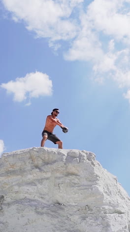 shirtless athlete lifting kettlebell. healthy sportsman is swinging kettlebell with two hands on the top of white hill. view from below on bodybuilder under blue sky. vertical video