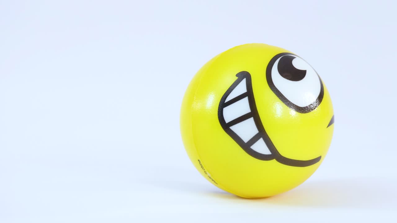 Una palla di gomma gialla con faccina sorridente rimbalza e rotola su una superficie bianca e pulita.
