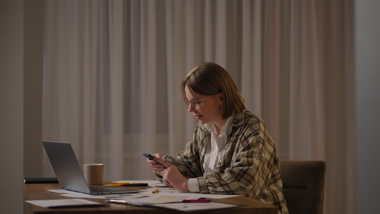 mujer feliz usando teléfono móvil mientras trabaja en casa con computadora portátil. mujer fresca sonriente con gafas enviando mensajes con teléfono inteligente. mujer hermosa y elegante navegando por el sitio en el teléfono celular