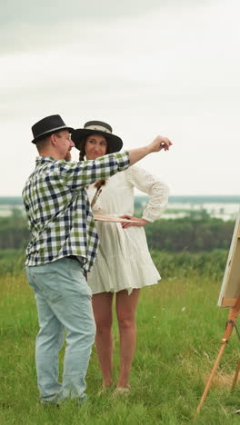 a man in a black hat and plaid shirt stands close to a woman in a white dress in a grassy field. they are engaged in a discussion or observation, with a drawing board set up in front of them