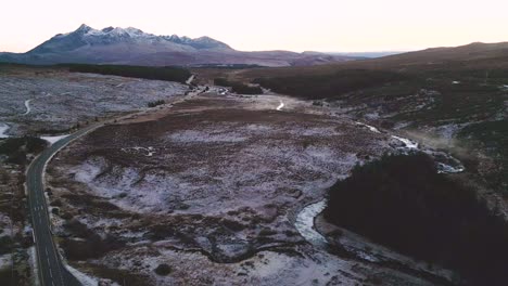 Camino-Sinuoso-A-Través-De-Un-Paisaje-Cubierto-De-Nieve-Con-Un-Telón-De-Fondo-Montañoso-Al-Atardecer,-Vista-Aérea