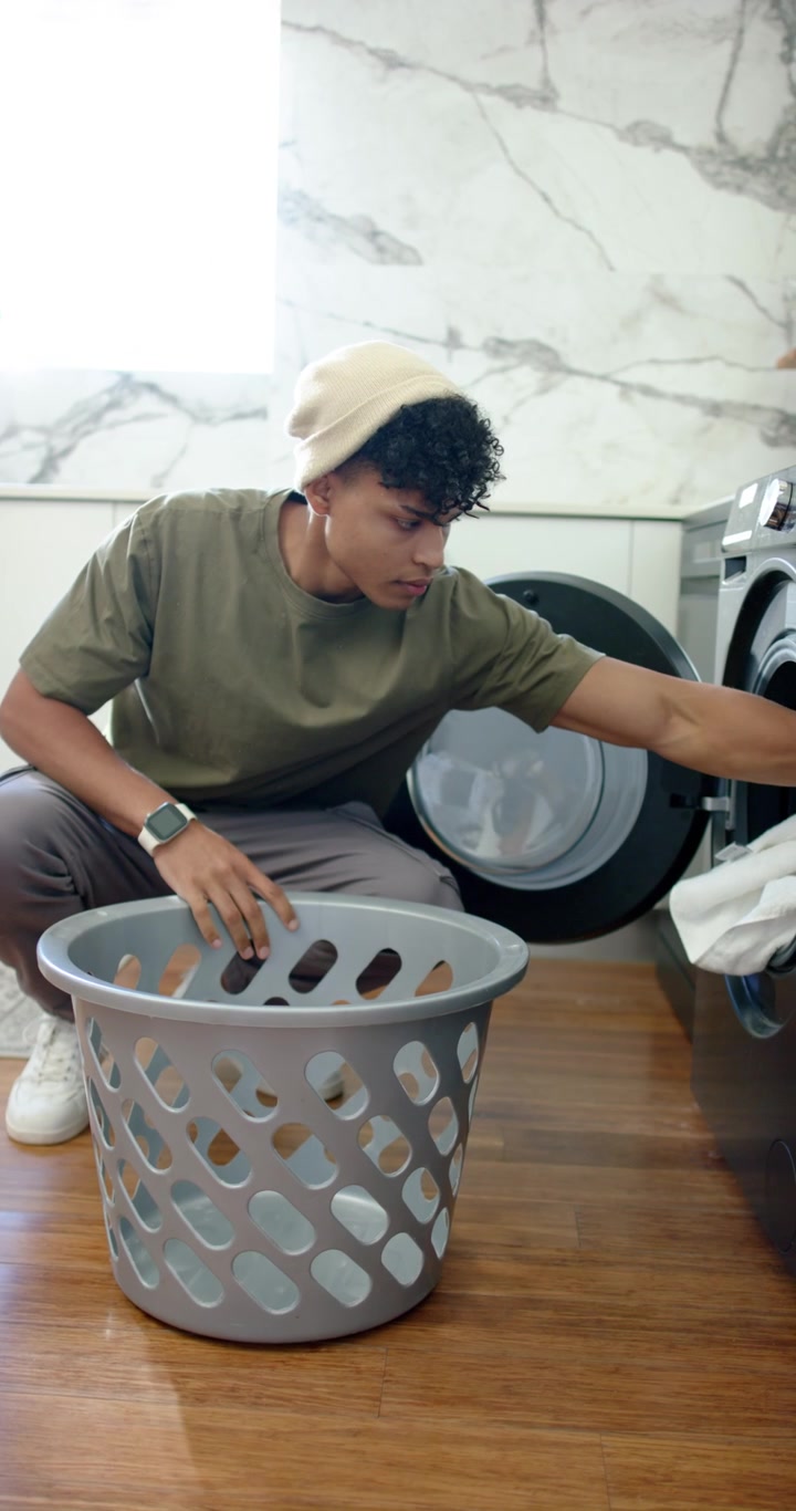 Vertical video: African American man at washer, searching basket, loading whites into drum to wash
