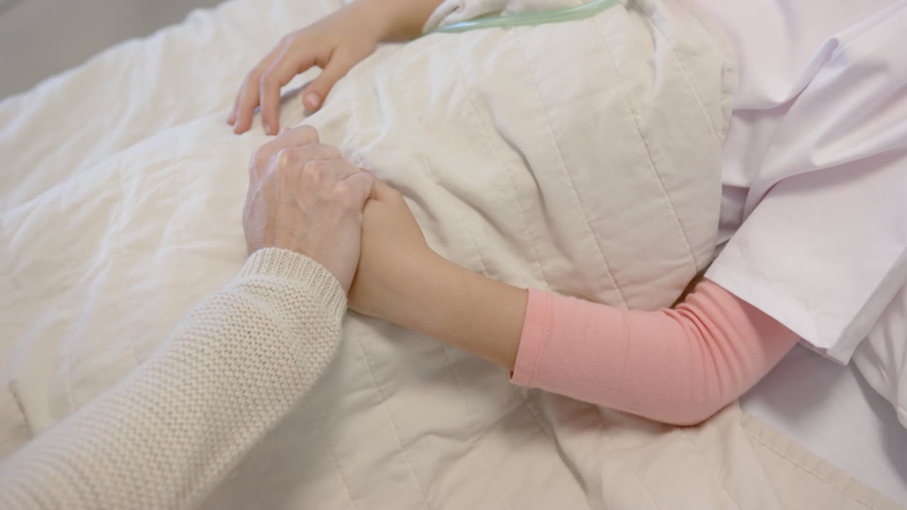la mano de la madre caucásica sosteniendo la mano de la hija paciente acostada en la cama del hospital, cámara lenta