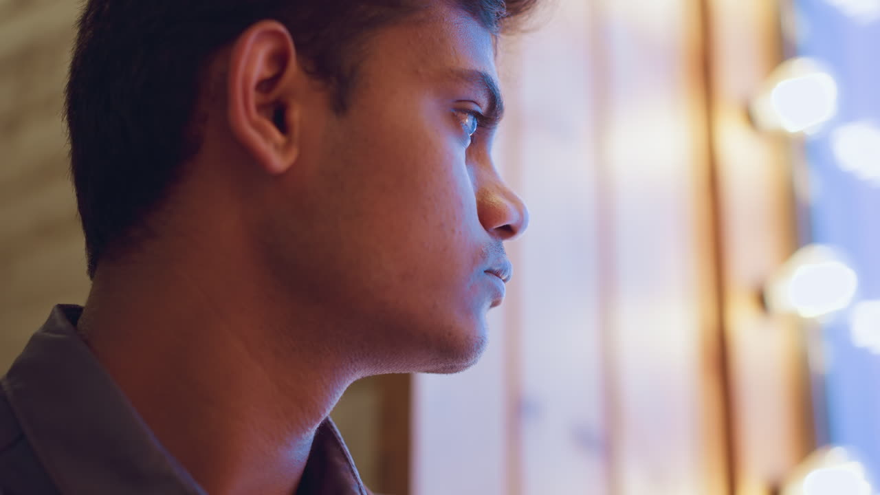 Close-up side profile of young man indoors gazing through softly lit window, eyes reflecting glow of ambient lights, creating serene contemplative atmosphere with sense of quiet emotion