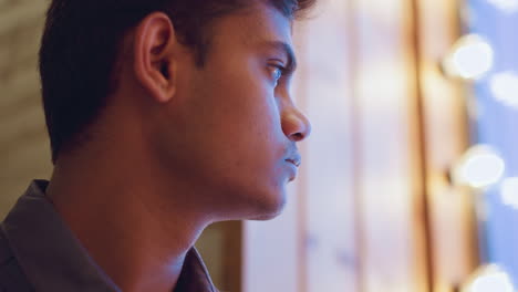 close-up side profile of young man indoors gazing through softly lit window, eyes reflecting glow of ambient lights, creating serene contemplative atmosphere with sense of quiet emotion