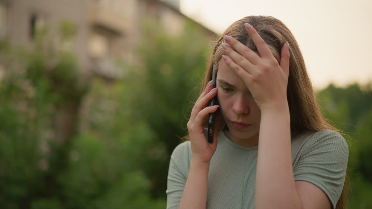 Young lady appears visibly frustrated and exhausted during phone call, holding her hand to her forehead in a gesture of stress, blurred background shows greenery and distant building
