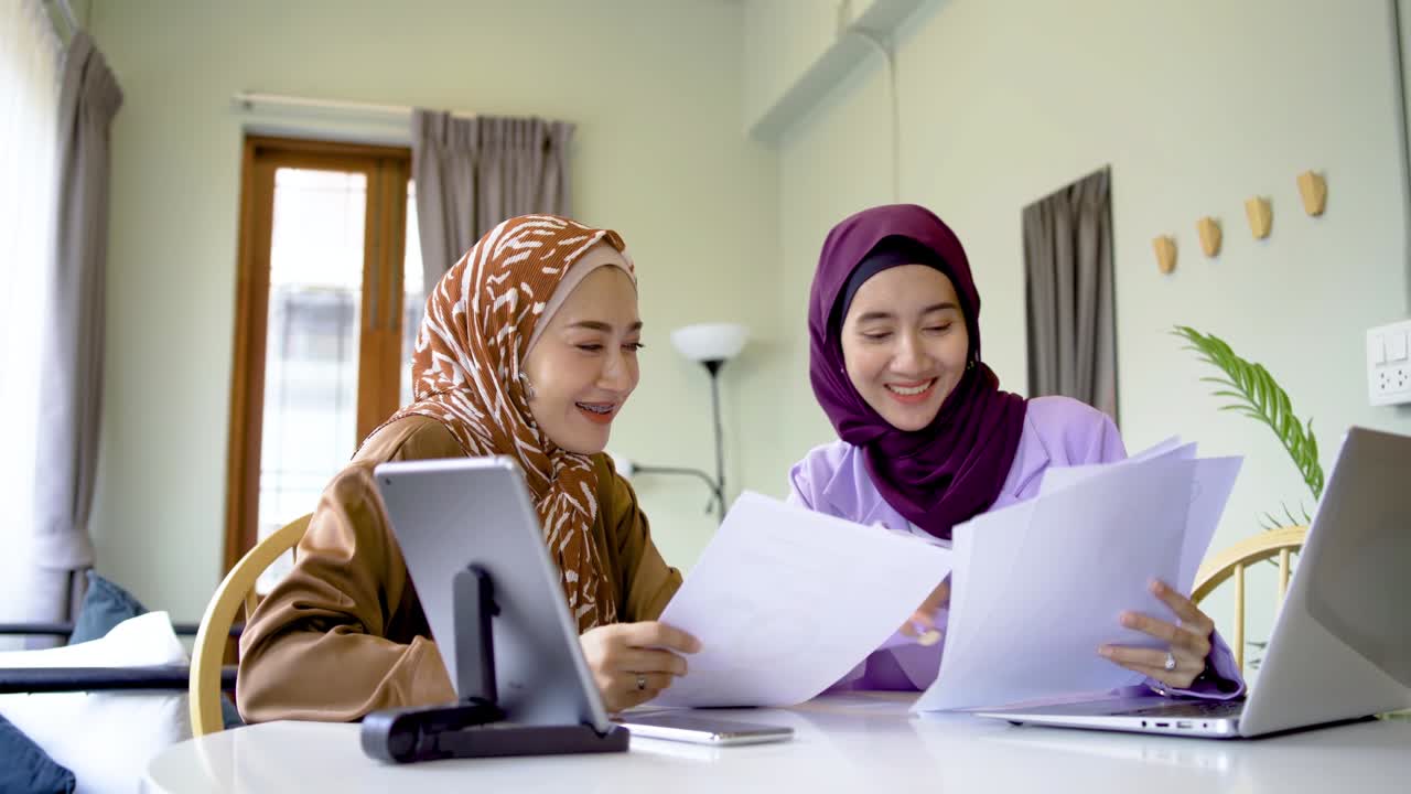 두 명의 히자브를 입은 무슬림 여성이 집에서 거실에서 그래픽 차트를 보여주는 노트북, pc, 태블릿 및 종이를 사용하여 함께 일하며, 서로에게 하이 파이브를 주고, 예상대로 완벽하게 작업을 완료했습니다.