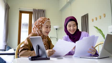 two muslim women wearing hijab working together with using laptop pc tablet and paper showing graphic chart in living room at home, give each other hi five, completed the work perfectly, as expected