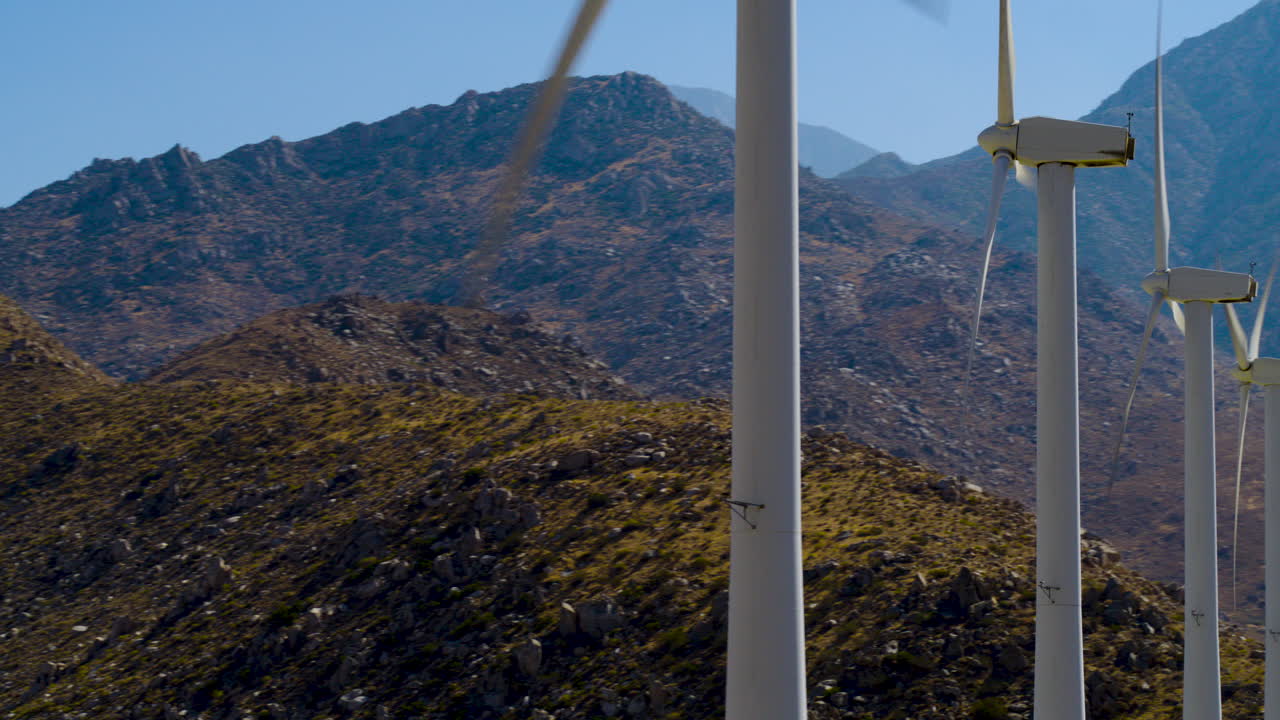Palm Springs backside of windmills spinning with the rugged San Jacinto Mountains rising behind them in California under a clear blue sky on a bright, sunny day. Aerial shot spinning around them