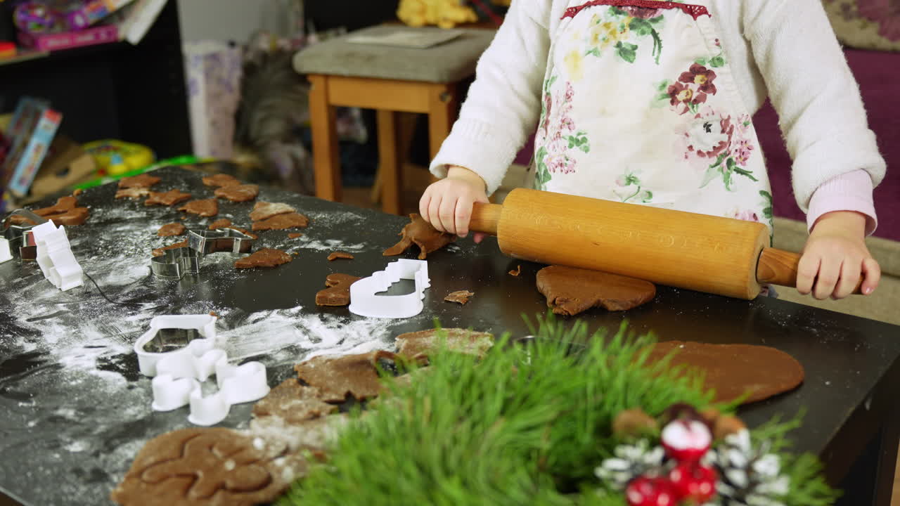 Hands press a rolling pin over gingerbread dough as cookie cutters and flour scatter the table, while festive greens and tiny Christmas ornaments decorate the corner. Close-up shot, static camera.