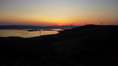 aerial drone view of wind turbines spinning on hills during colorful sunset in romania, with lakes and silhouettes.