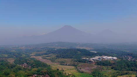 Silhouette-of-twin-mountain-peeks-in-far-distance-behind-misty-area,-aerial-drone-view