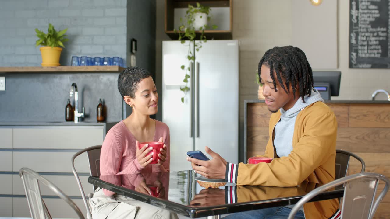 Diverse couple sharing smartphone screen after smiling and holding red mugs at home kitchen table