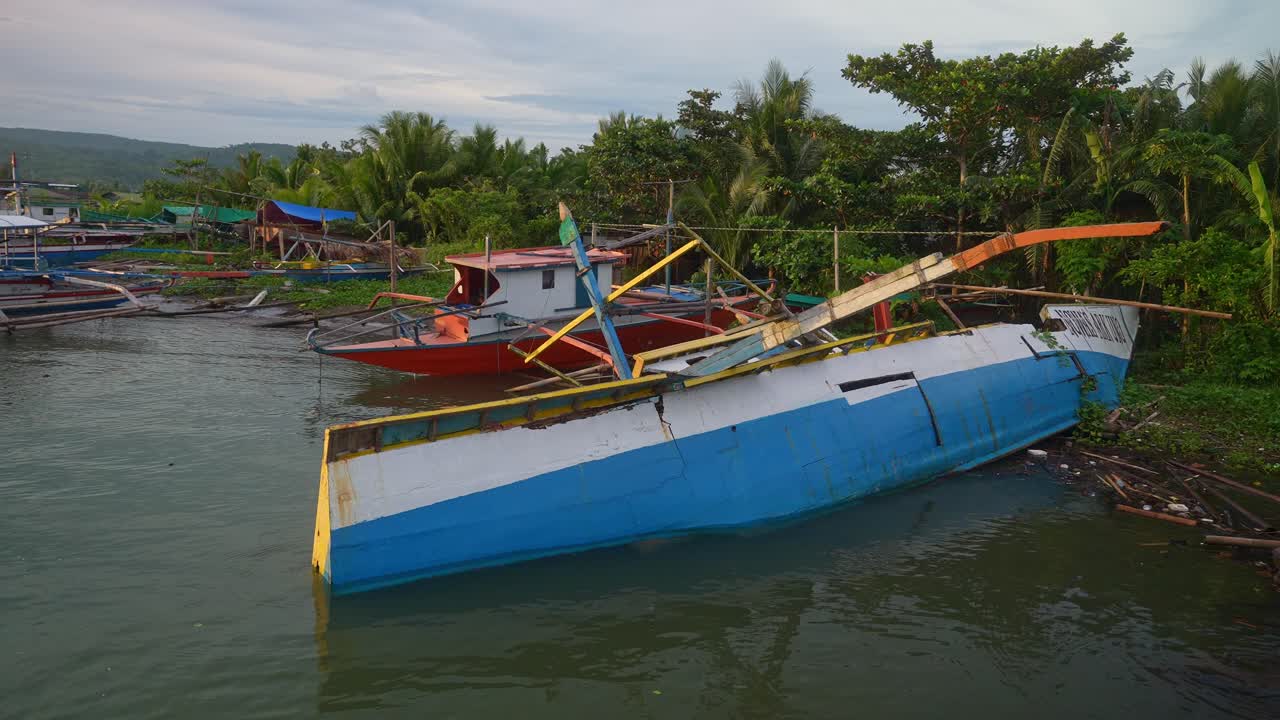 A fixed shot of a damaged blue and white outrigger boat tilted in shallow water near the tropical shore of Mauban Port, Quezon Province Philippines with palm trees under an overcast sky