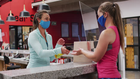 Two-caucasian-women-wearing-face-masks-talking-at-reception-of-gym-and-disinfecting-hands