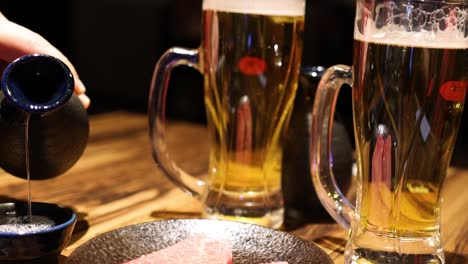 a person pours sauce into a small dish next to a beer mug and raw steak on a wooden table.