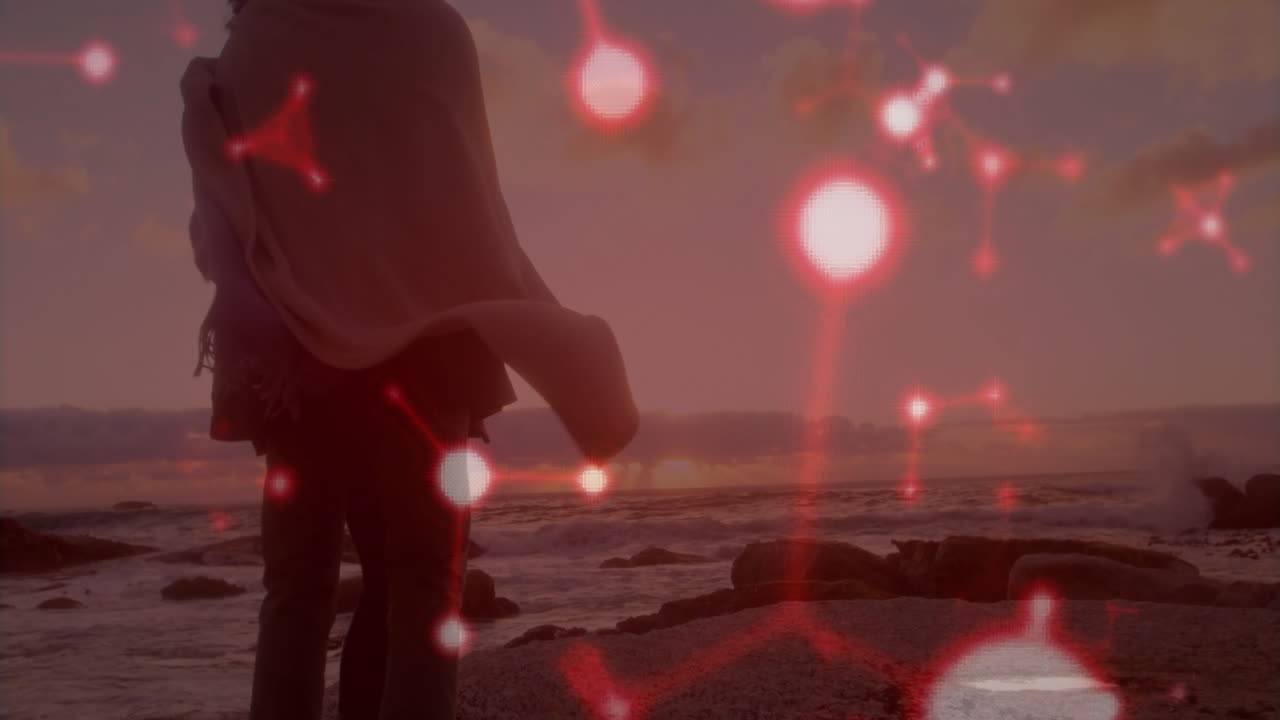 woman and man standing on rocky beach at sunset, showing floating red network nodes for technology