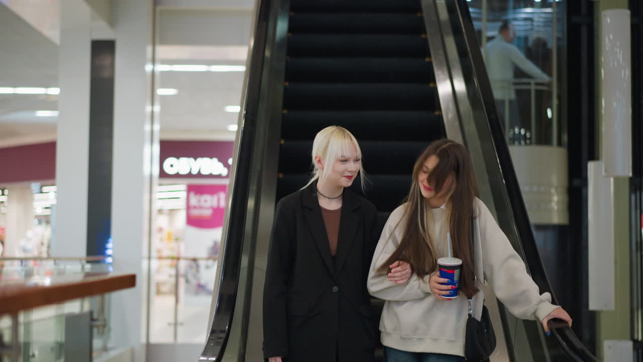 Girls step out from escalator in shopping mall holding drink, smiling with relaxed expressions, enjoying leisure time together in modern retail environment surrounded by shops