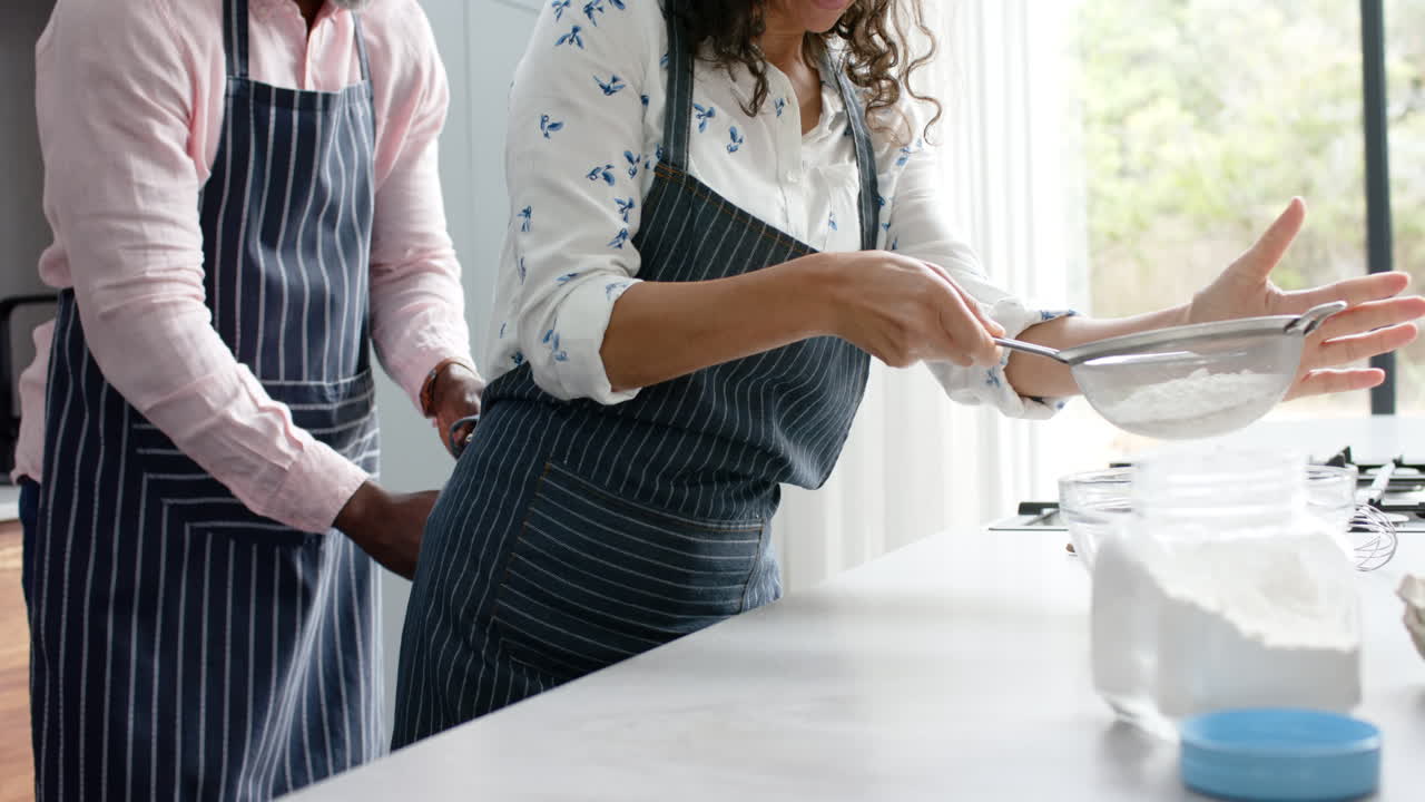 pareja biracial feliz usando delantales y horneando en la cocina, cámara lenta