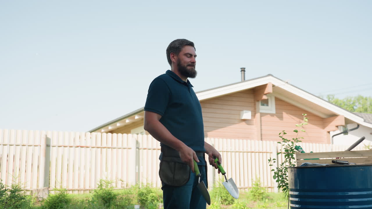 Young gardener tosses trowel, catches, slides tool into apron belt on waist, then removes again, standing near blue drum and wooden crate in sunny backyard