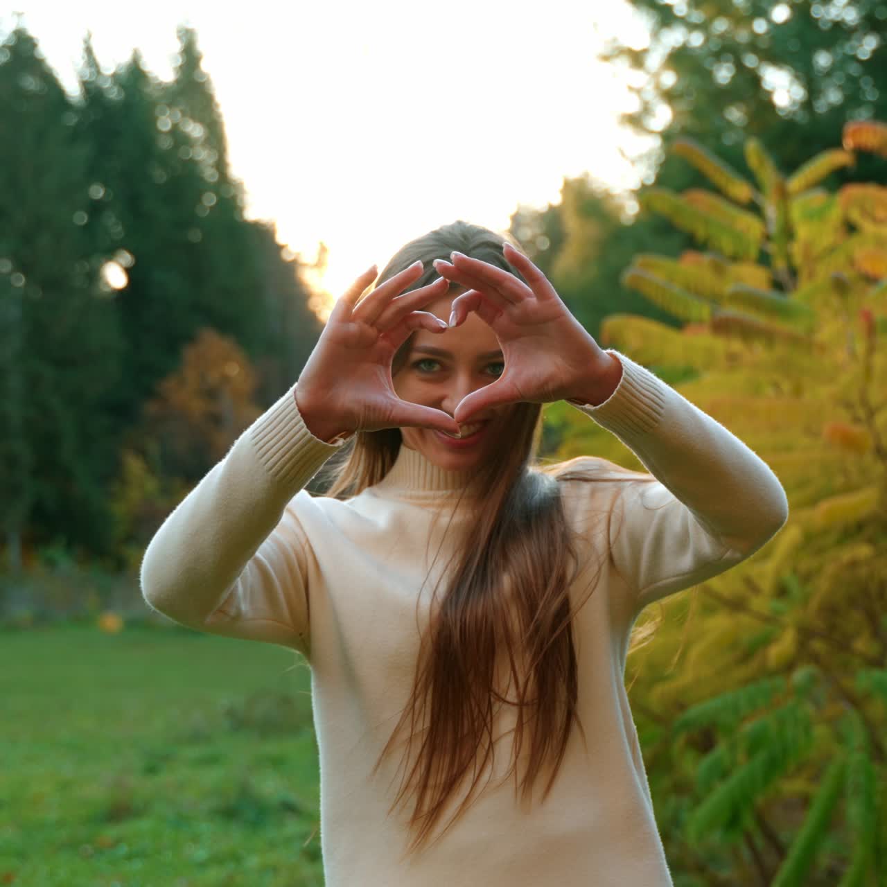 Happy beautiful woman in white warm sweater outdoors. Lady smiling to the camera and showing heart sign to the camera. Autumn nature at backdrop