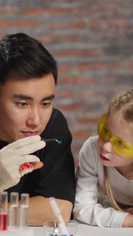 young man asian scientist and concentrated little girl student examine small crystal in tongs sitting at table at fun science lesson in laboratory