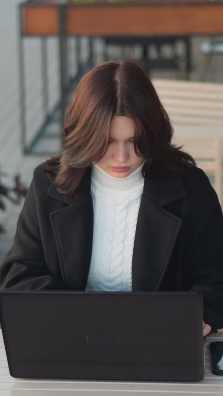 Lady focused on laptop outdoors in urban setting with her phone on the table, wearing stylish black coat, decorative black pillar nearby, flower vessel near by and chairs in background