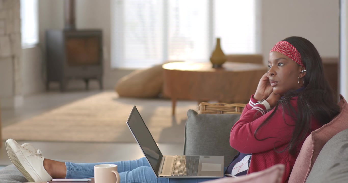 African American woman checking laptop, inserting earbud for audio and adjusting on sofa