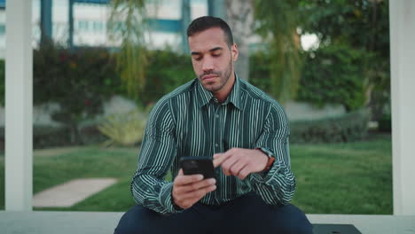 Young-man-checking-smartphone-sitting-in-bench-outdoors.