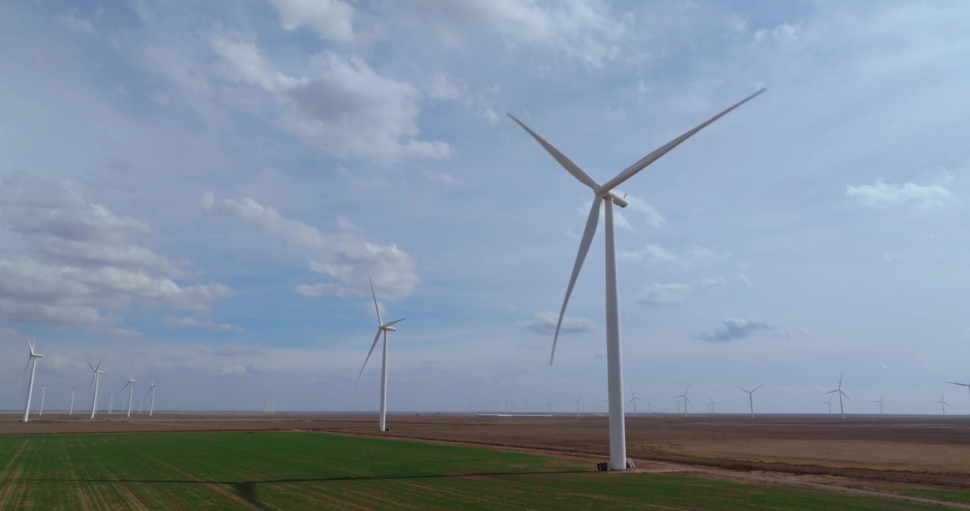 Un drone aereo avanza verso grandi turbine eoliche rotanti che si ergono sopra le pianure piatte del Texas con campi coltivati verdi e un cielo nuvoloso e drammatico sopra.