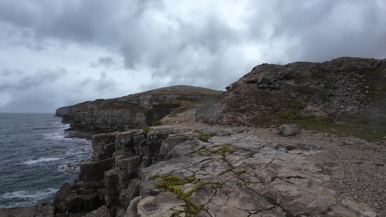 Dramatic aerial footage captures rugged rock formations and crashing waves on a moody, overcast day at the Isle of Purbeck, Dorset. The wide ocean view creates a serene yet powerful atmosphere