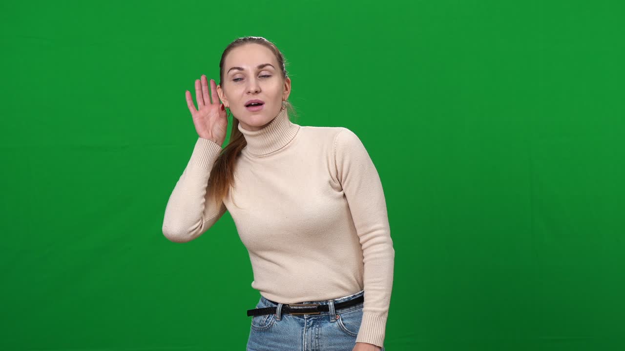Adorabile donna caucasica bruna che ascolta qualcosa tenendo la mano all'orecchio e annuendo. Ritratto di una signora che origlia su sfondo verde chromakey. Concetto di femminilità e pettegolezzo.