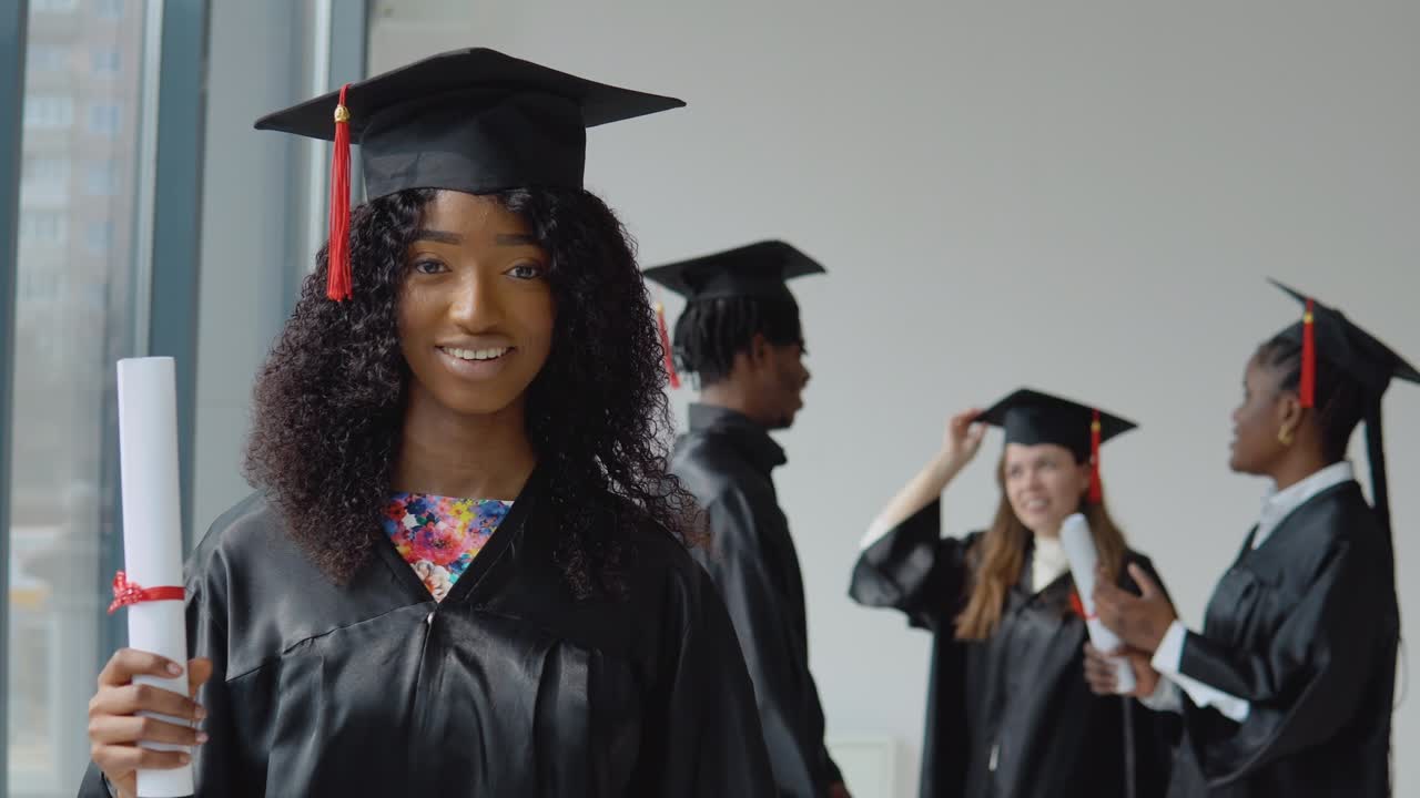 una joven afroamericana está de pie en primer plano y mira a la cámara con un diploma en la mano. un joven afroamericano y una mujer y una mujer caucásica se comunican detrás.