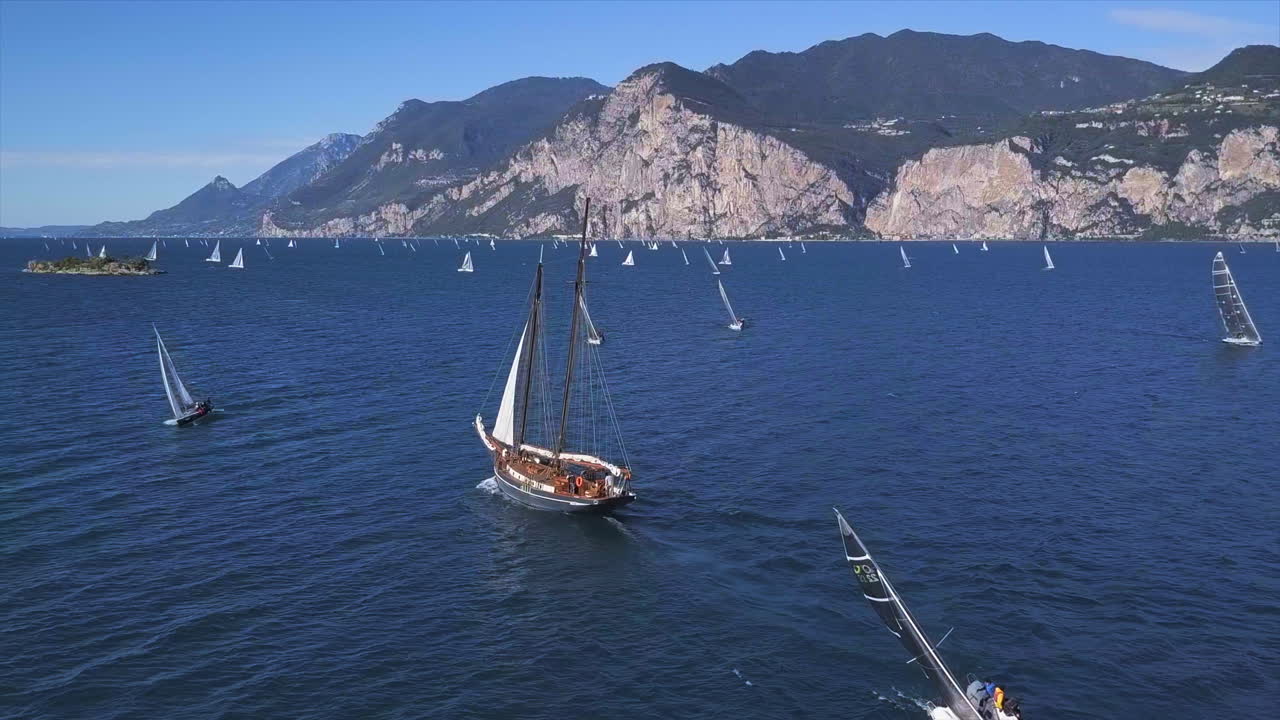 Aerial shot of a historic sailing ship on the Lago di Garda with the coastline in the background on a bright sunny day. A smaller sailboat is crossing the foreground