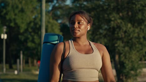 plus size african-american woman walking with exercise mat through the park in a summer day.