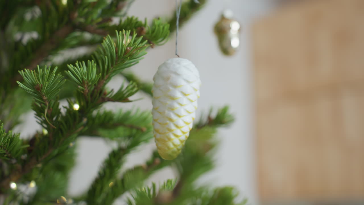 Beautiful white ornament hanging on Christmas tree surrounded by vibrant green pine branches and warm glowing lights, cozy festive holiday setting captured with soft background