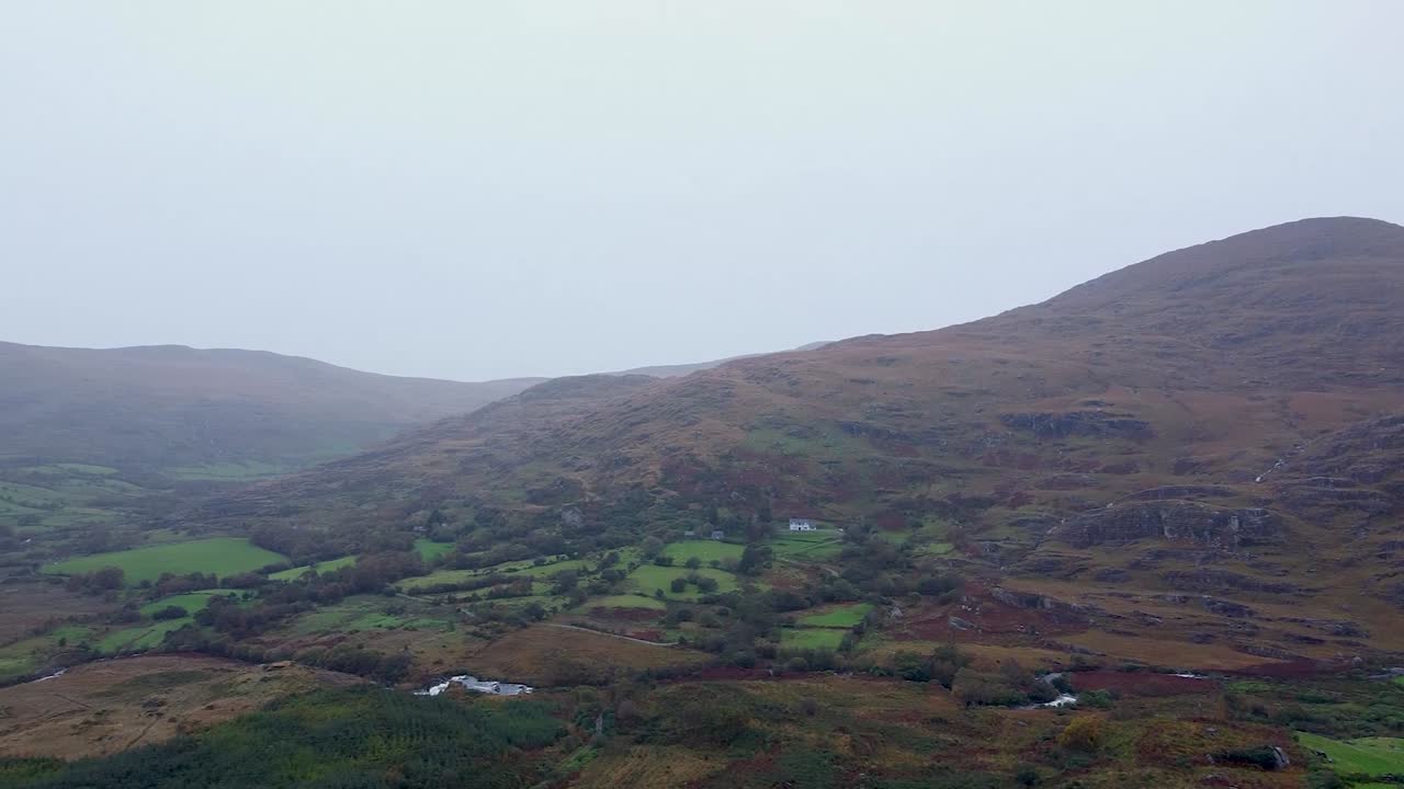 Panoramica di apertura che rivela la vasta e aspra bellezza della campagna rurale irlandese, con campi verdi a mosaico nella valle di montagna sotto un cielo coperto.