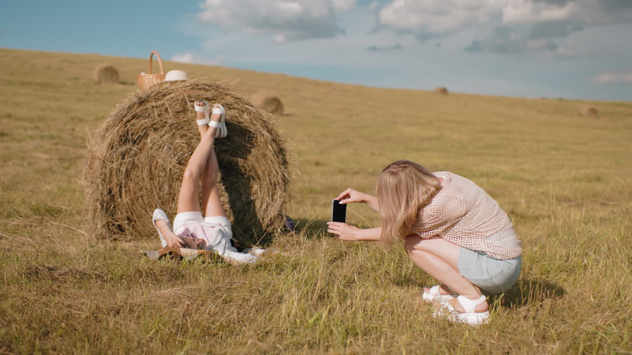Вид сзади на женщину, фотографирующую подругу, лежащую на тюке сена с поднятыми ногами, наслаждающуюся природой, корзина для пикника и шляпа лежат на сене, пока золотистое поле простирается под ярким небом