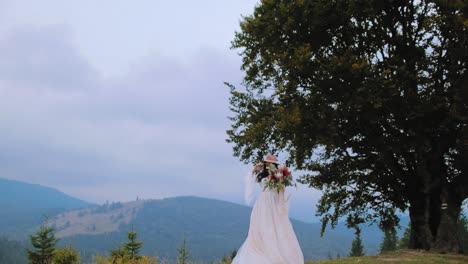 Bride in white dress near the green tree. Beautiful woman in long white dress standing with bouquet of flowers against mountainous area.