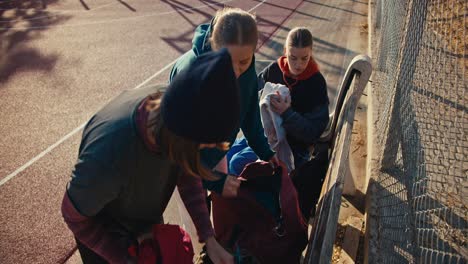 A-trio-of-girls-in-sportswear-put-their-things-in-a-backpack-on-a-street-sports-field-after-playing-basketball-on-a-sunny-summer-morning.-Three-blonde-girls-in-sports-summer-uniforms-collect-their-things-after-playing-basketball