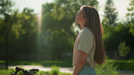 cyclist leans back with head slightly tilted, savoring warm sunlight as her hand rests on bicycle handlebars, soft-focus greenery and trees