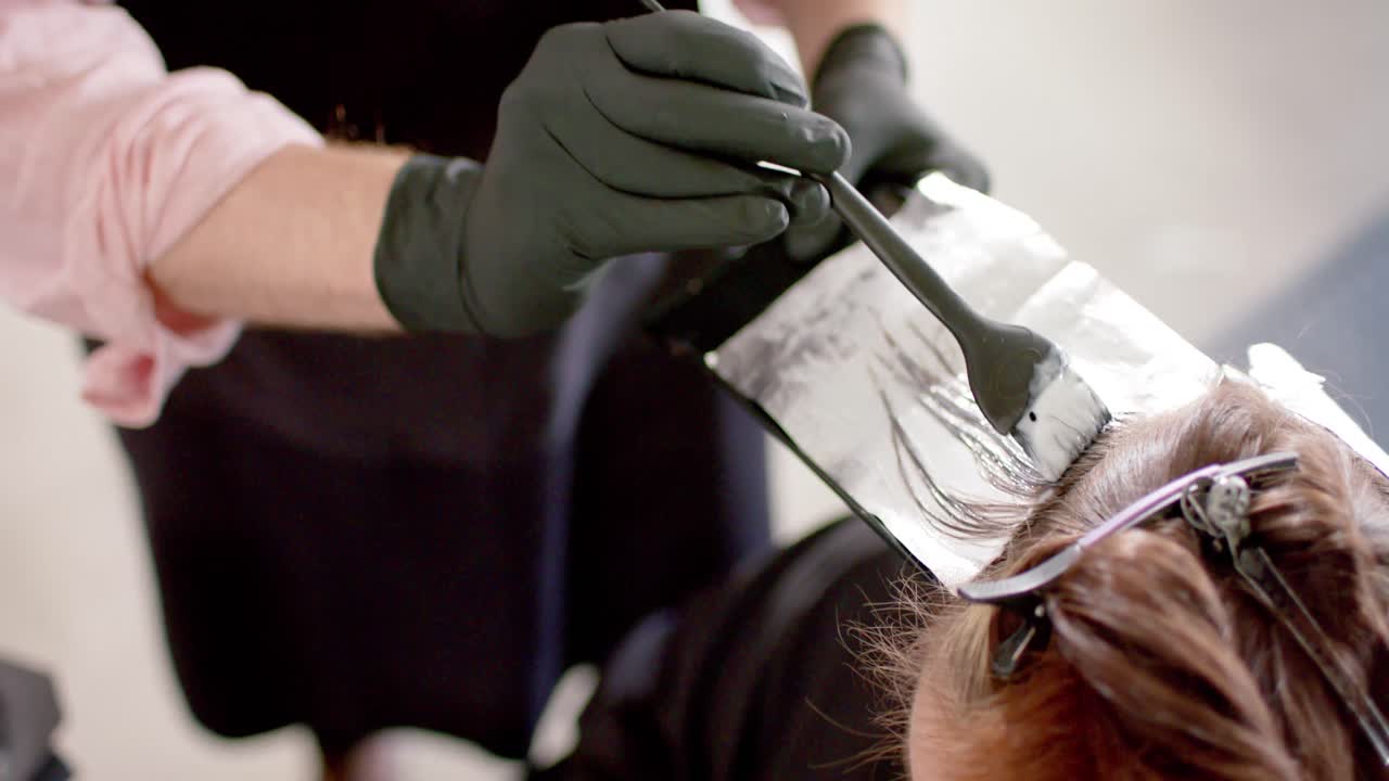 Caucasian male hairdresser highlighting client's hair with brush and foil at salon, in slow motion