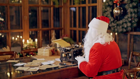 back view of santa claus sitting on the chair at and printing a letter on an old machine. santa's residence and the table with many envelopes written by father frost in the room.