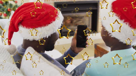 golden star icons falling against father and son having a videocall on smartphone during christmas