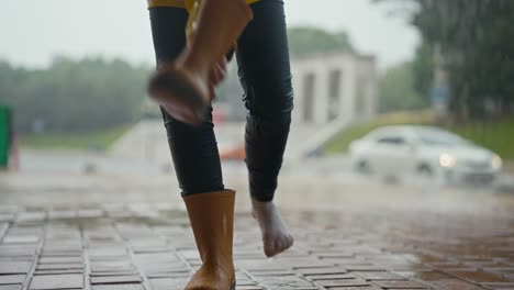 close-up of a teenage girl taking off her orange rubber boots and draining the water from them while walking and raining in the park