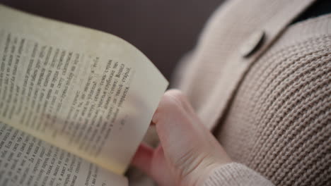 primer plano de una mujer con ropa tejida abriendo la siguiente página de un libro, el ambiente acogedor y relajado hace hincapié en la lectura tranquila con un enfoque en la textura suave de su prenda de punto